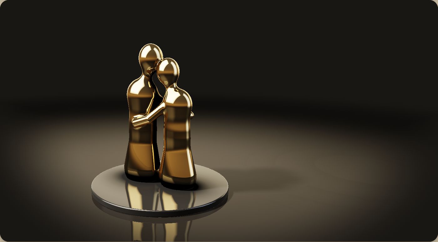 A polished gold sculpture of two figures leaning head to head, standing on a black plinth in a dark studio